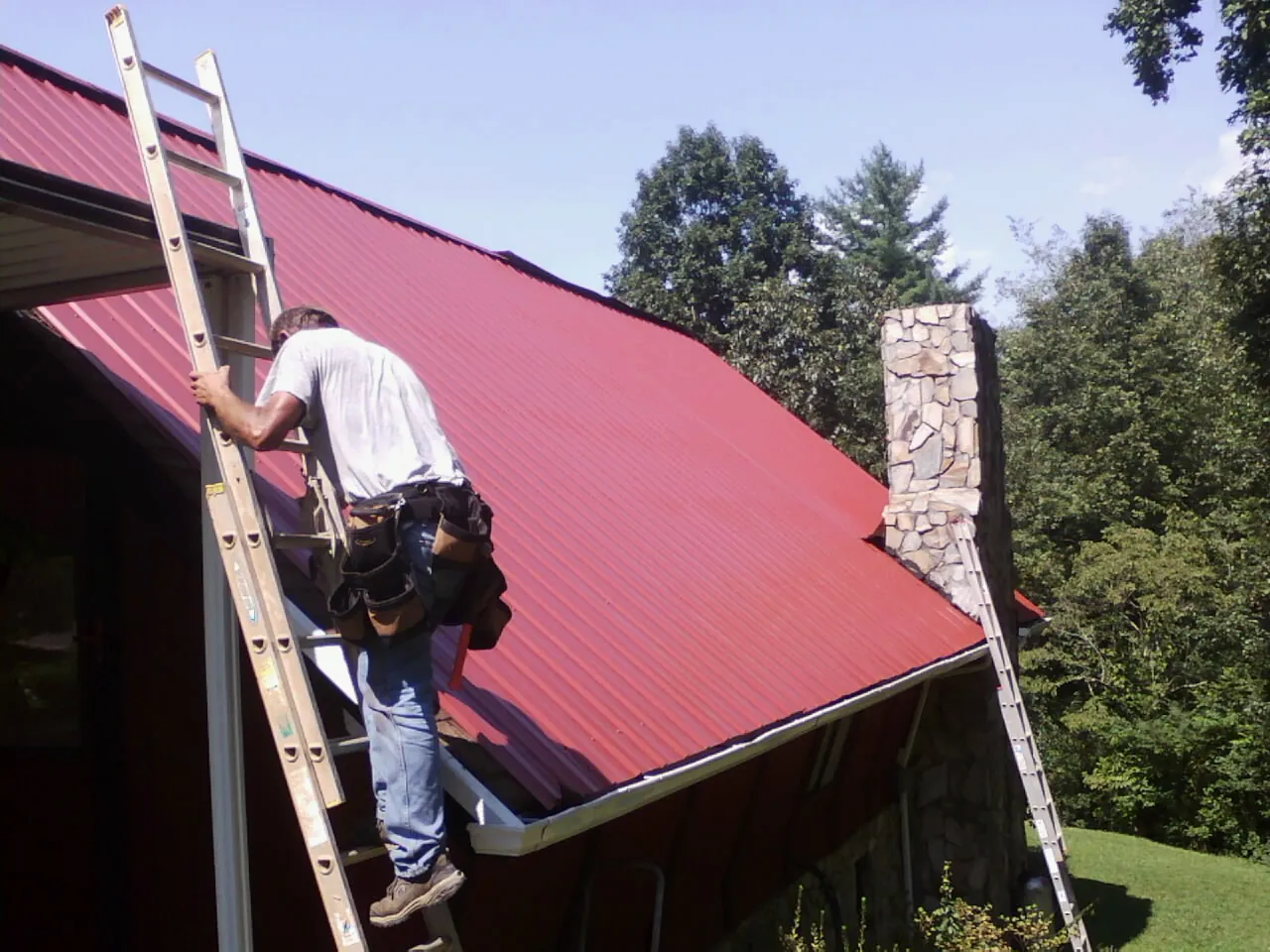 Roofer on a ladder beside a red metal roof and stone chimney — Soffit & Fascia Repair by Roofing Brothers in Danevang